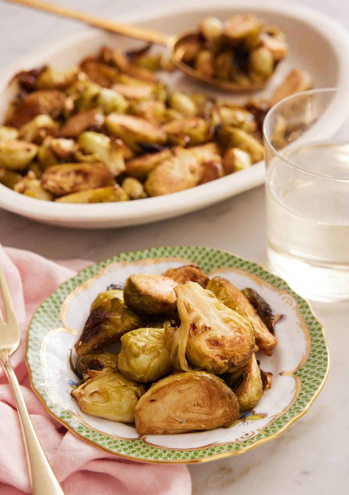 A bowl of air fryer Brussels sprouts with a drink and platter of more sprouts in the background.
