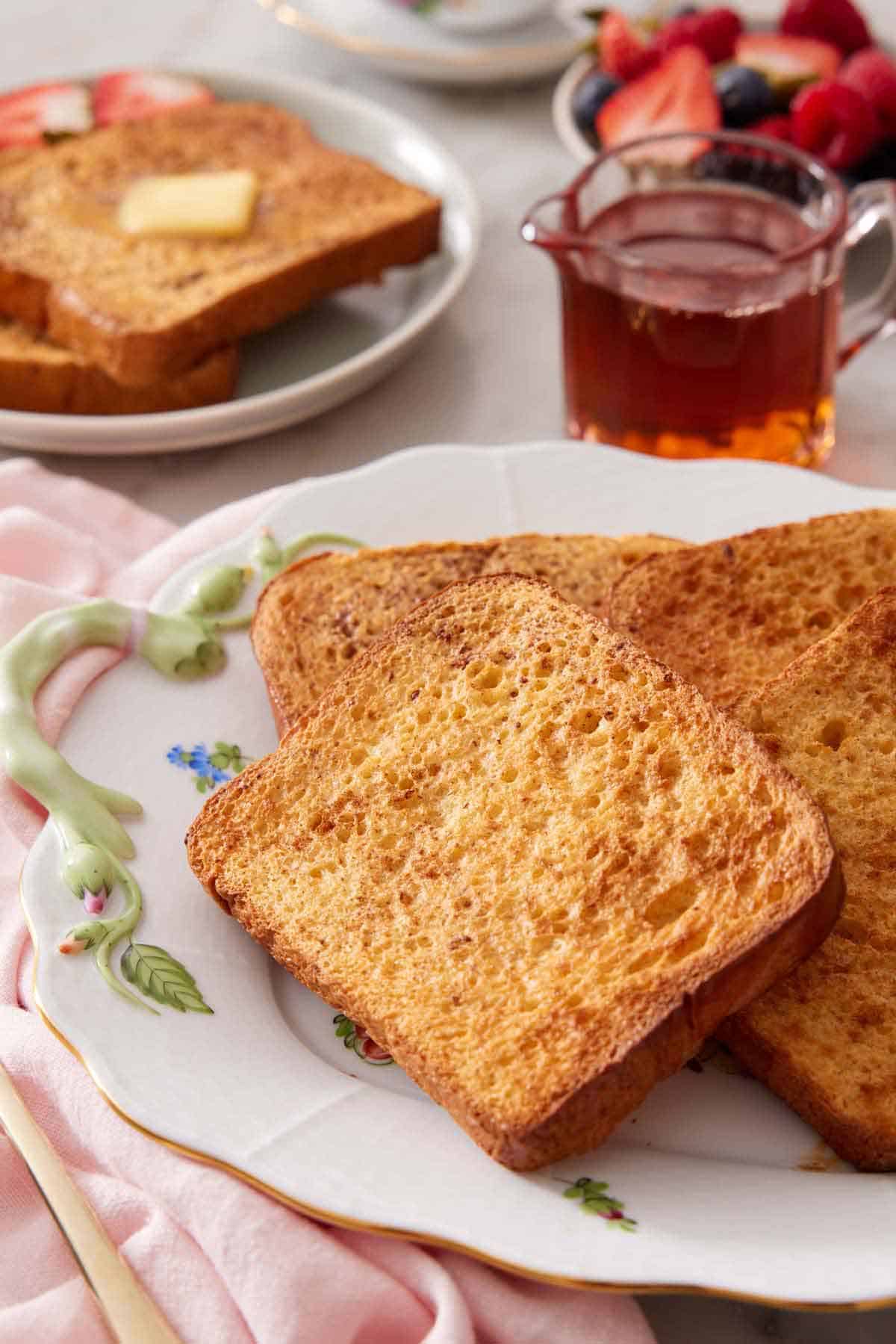 A platter with a pile of air fryer french toast with some maple syrup in the background along with a plated serving.
