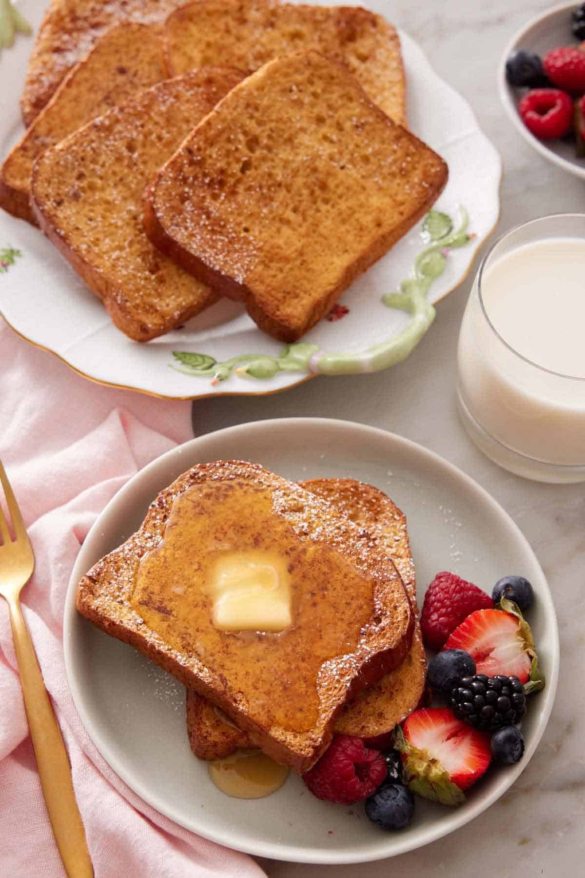 Overhead view of a plate of air fryer french toast with powdered sugar, syrup, butter, and berries. A platter of more and a glass of milk beside it.