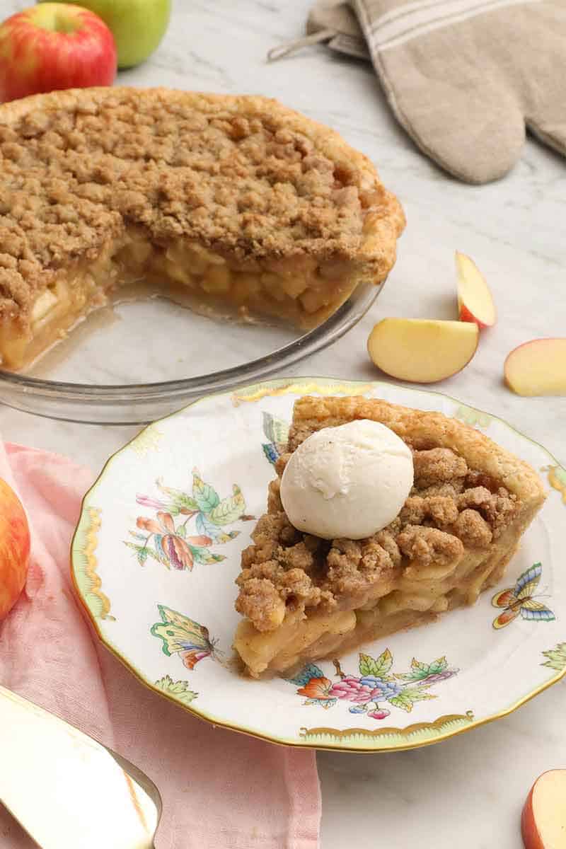A plate with a slice of apple crumble pie with ice cream on top and the cut pie in the background.