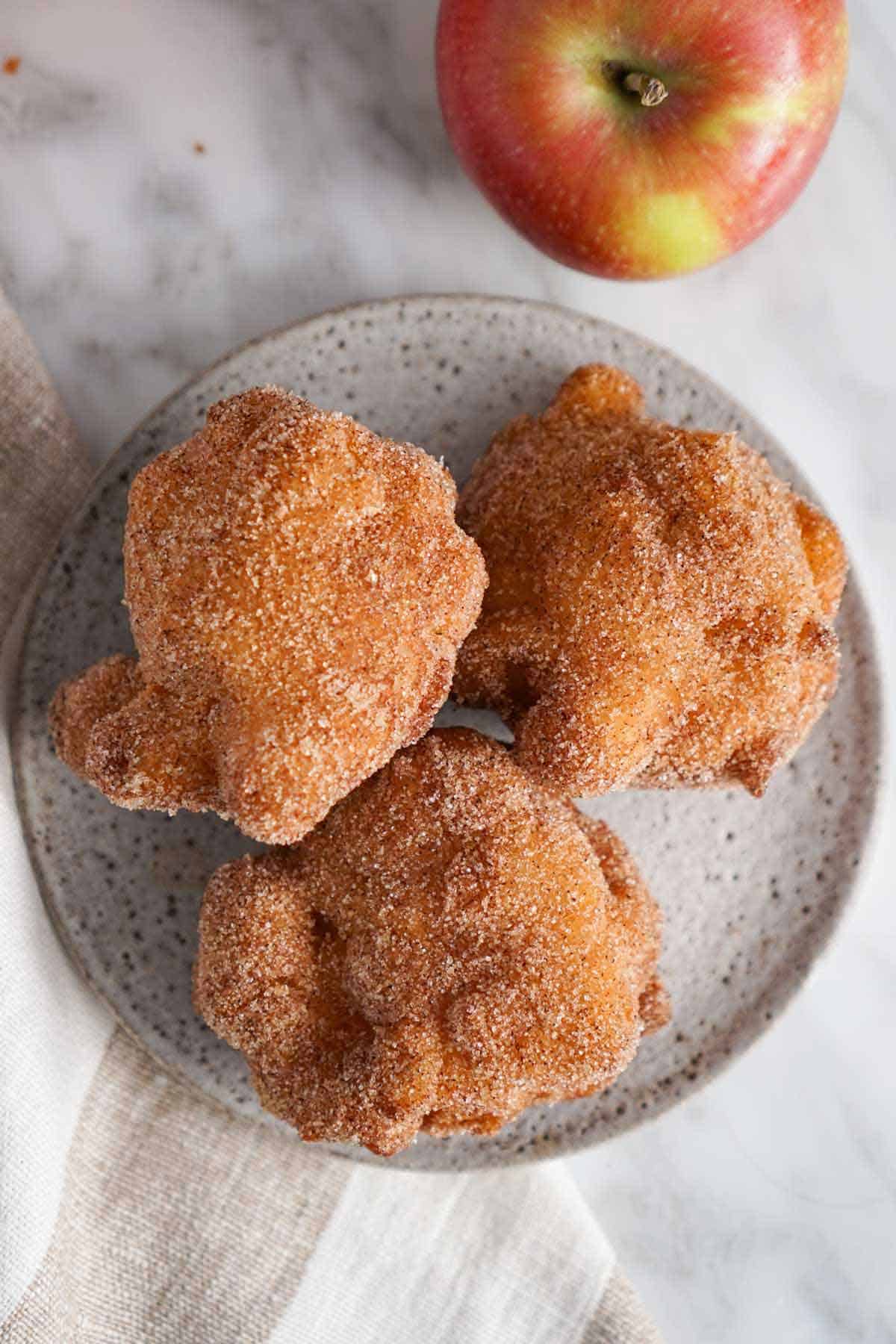 Overhead view of a plate with three apple fritters coated in cinnamon sugar. An apple beside it.