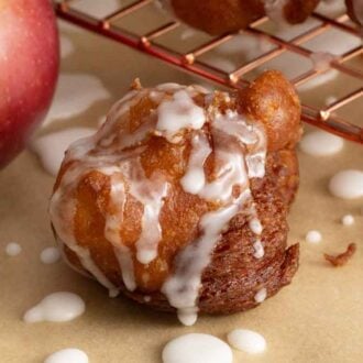 An apple fritter topped with a glaze with an apple and wire rack in the background.