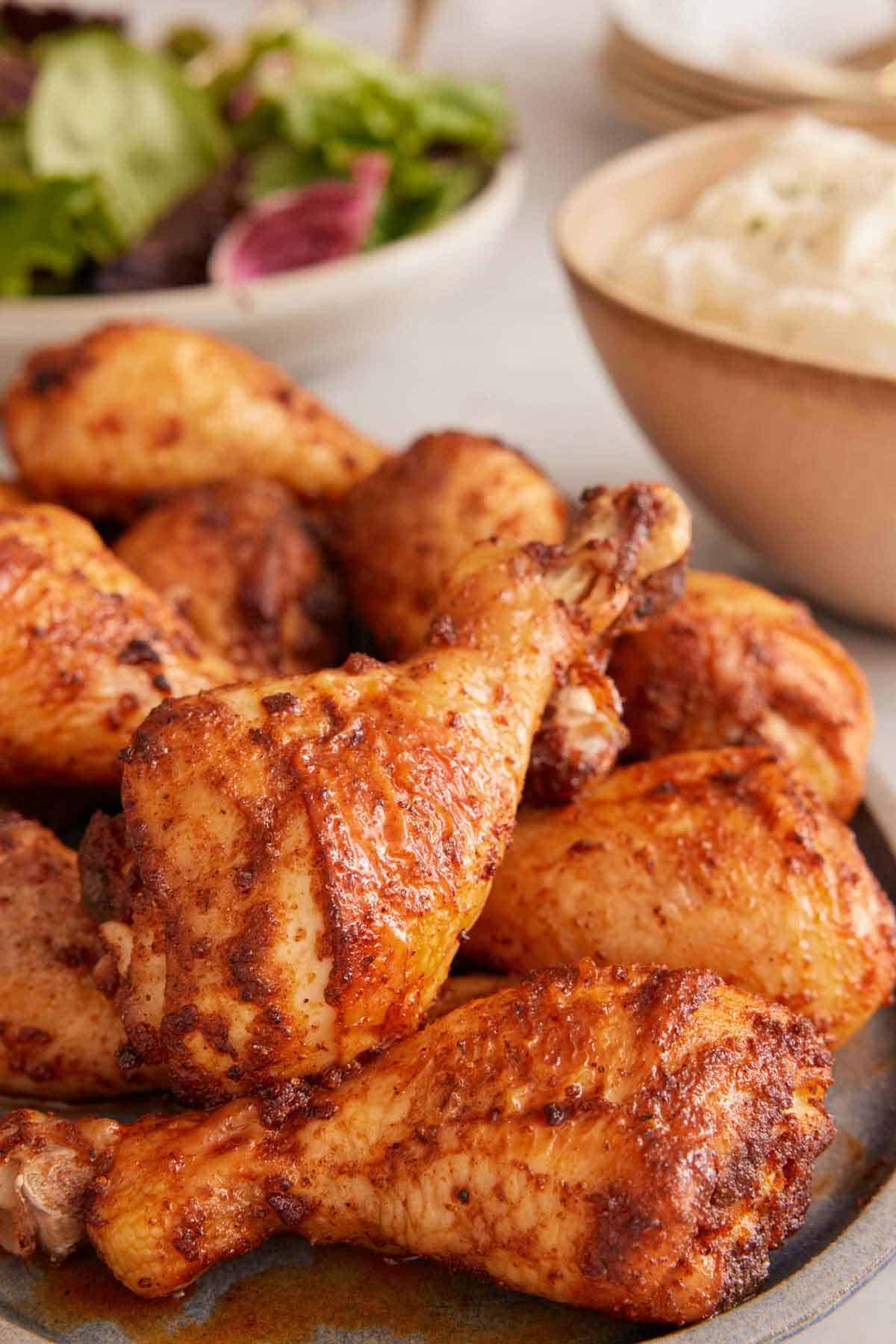 A small pile of baked chicken legs on a platter. Mashed potatoes and salad in the background.