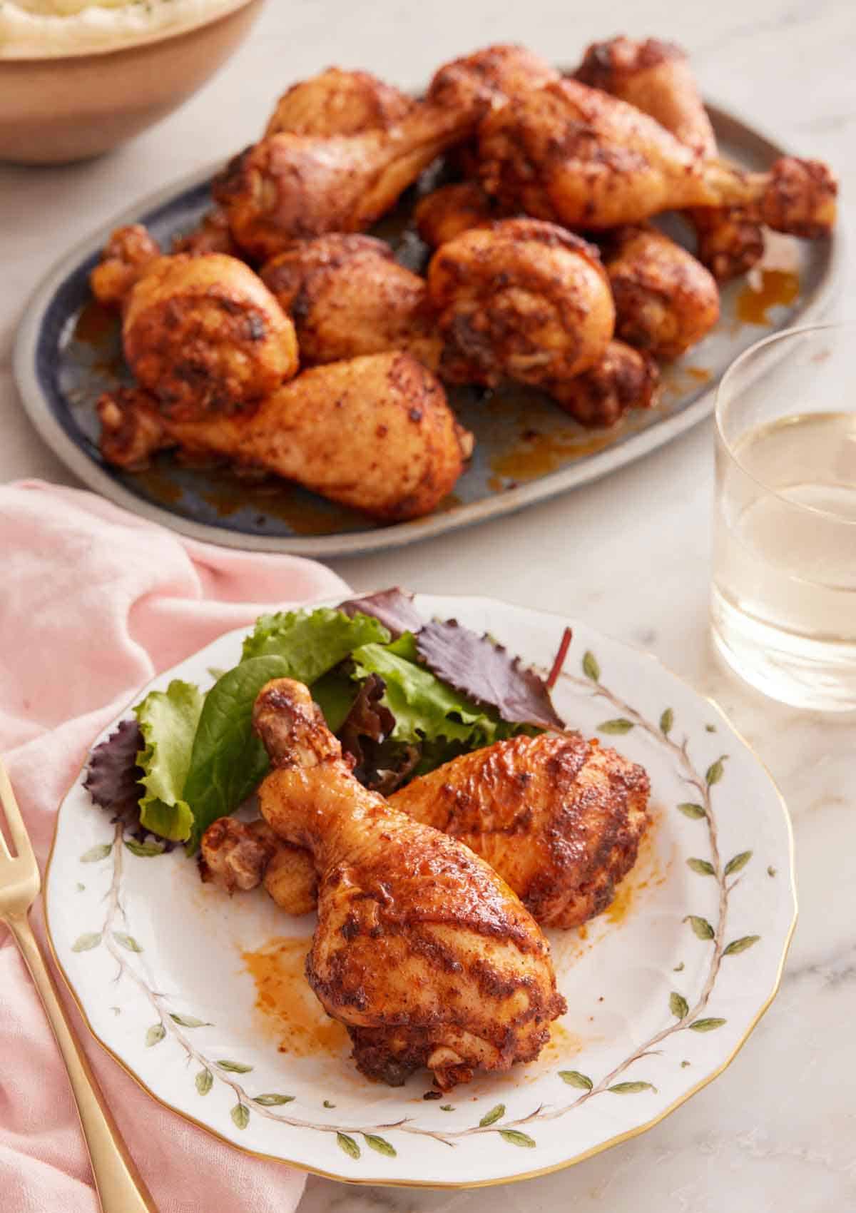 A plate with two baked chicken legs with some salad with a platter in the background along with wine.