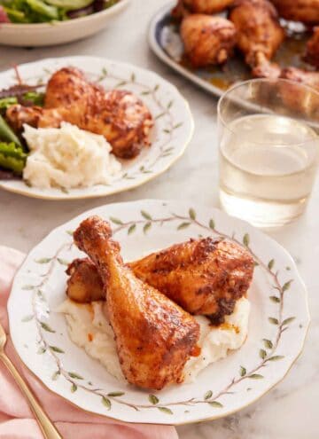 A plate with two baked chicken legs over mashed potatoes with a glass of wine and another plated serving in the background.