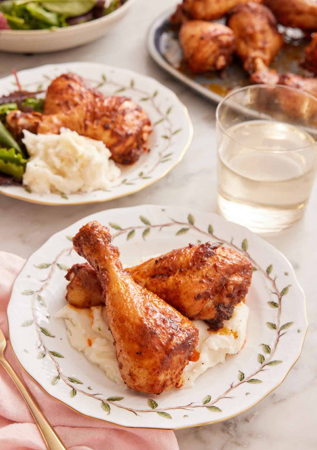 A plate with two baked chicken legs over mashed potatoes with a glass of wine and another plated serving in the background.
