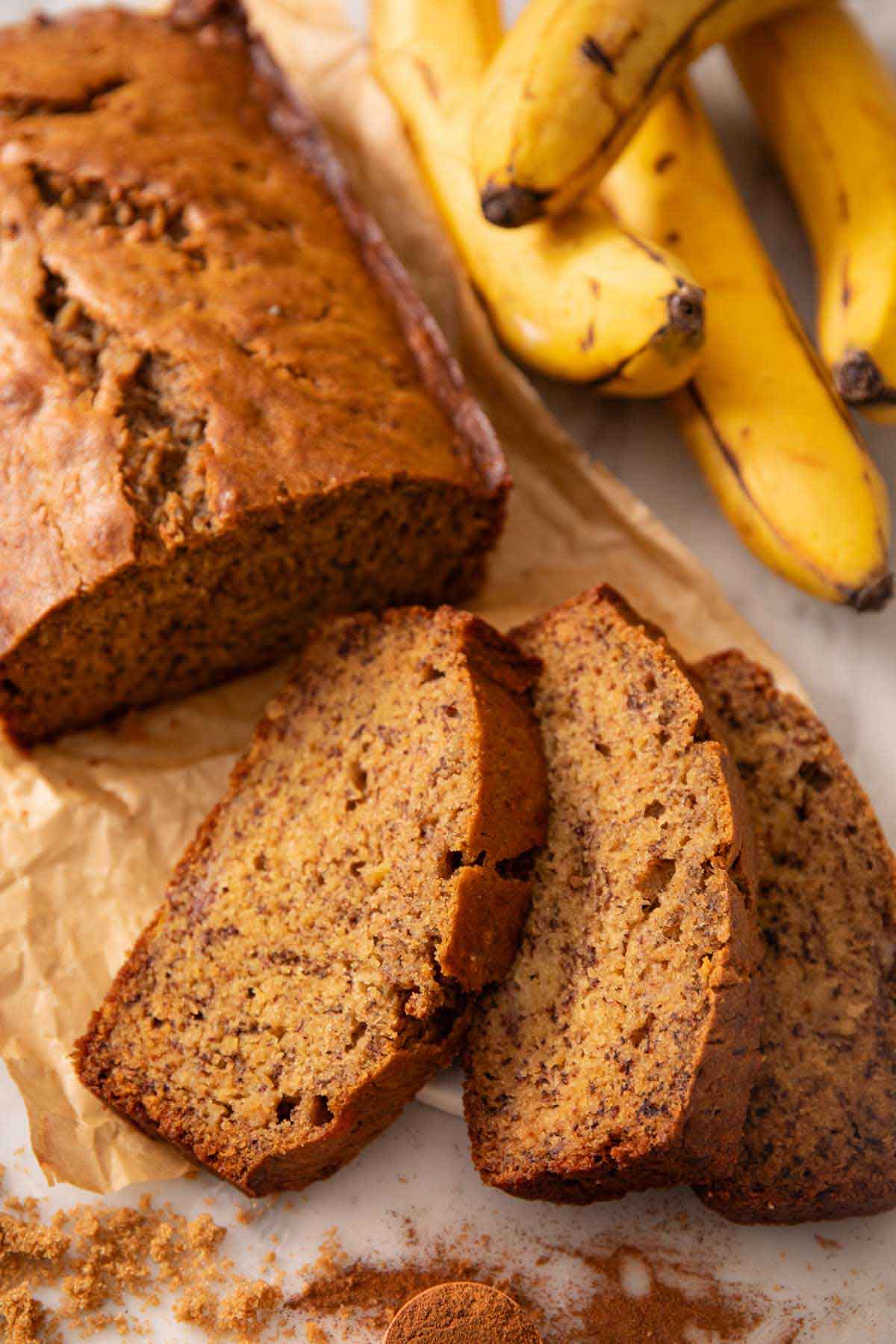 A loaf of banana bread with three slices in front. Bananas in the background.