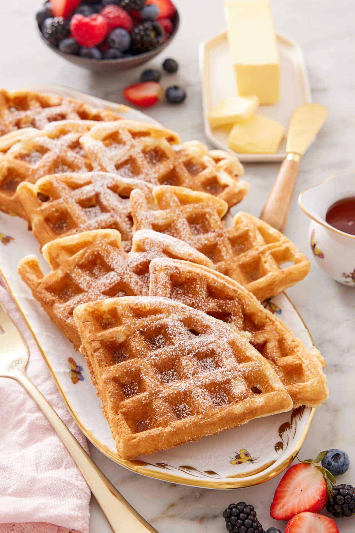 A platter with multiple triangle pieces of Belgian waffles topped with powdered sugar. Butter and berries in the background.