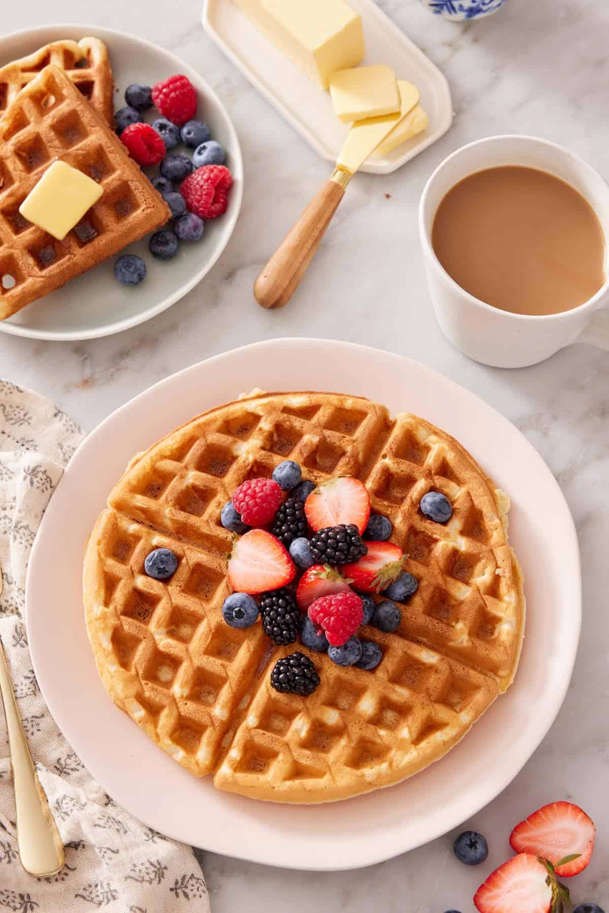 A plate with a Belgian waffle topped with berries. Coffee, butter, and more waffles in the background.