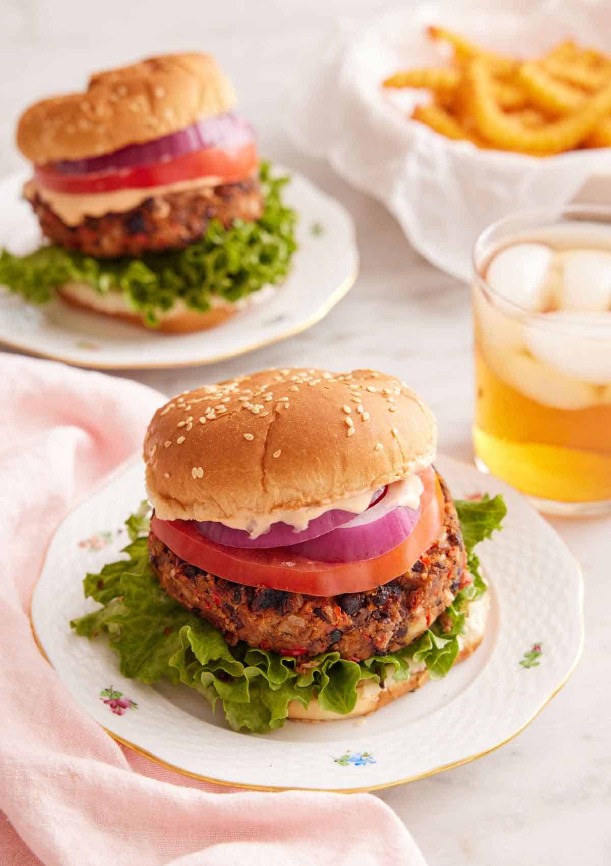 A plate with a black bean burger with a drink in the back along with a second burger and bowl of fries.