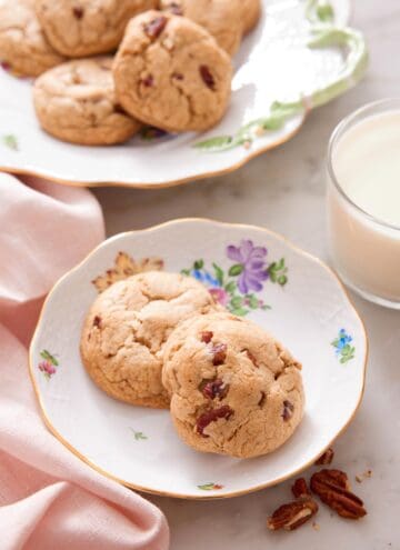 Two butter pecan cookies on a plate with a glass of milk and platter in the background.