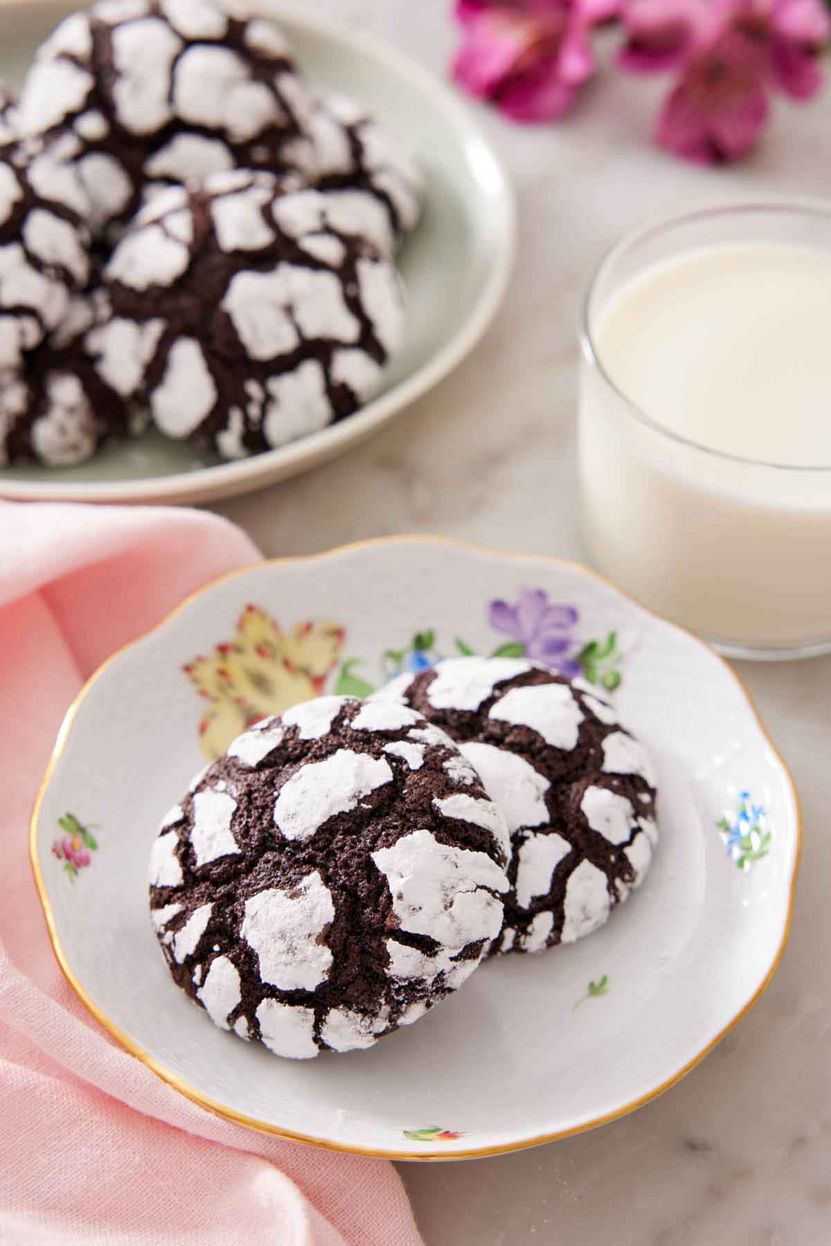 A plate with two chocolate crinkle cookies with a glass of milk and platter of more cookies in the background.