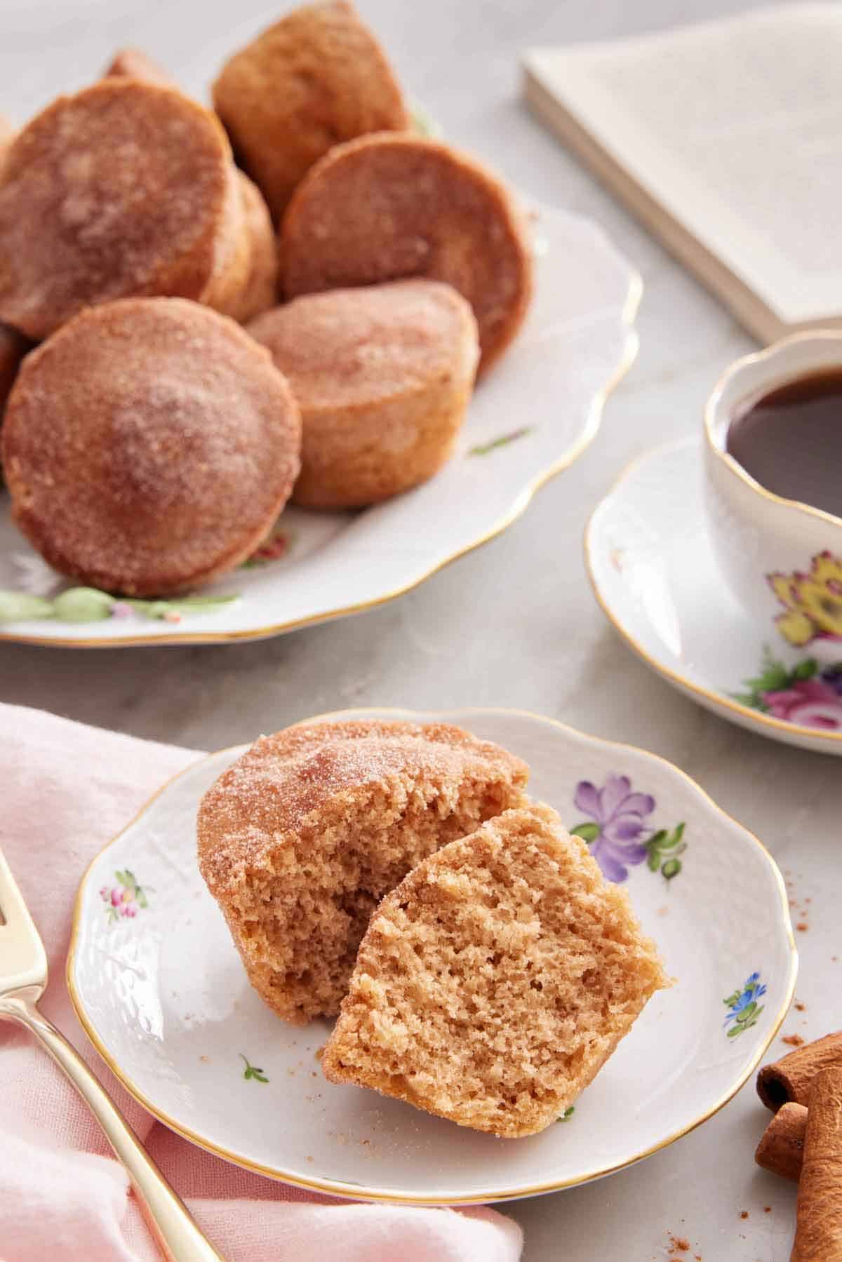 A plate with a cinnamon muffin cut in half with a platter in the background along with coffee and a book.