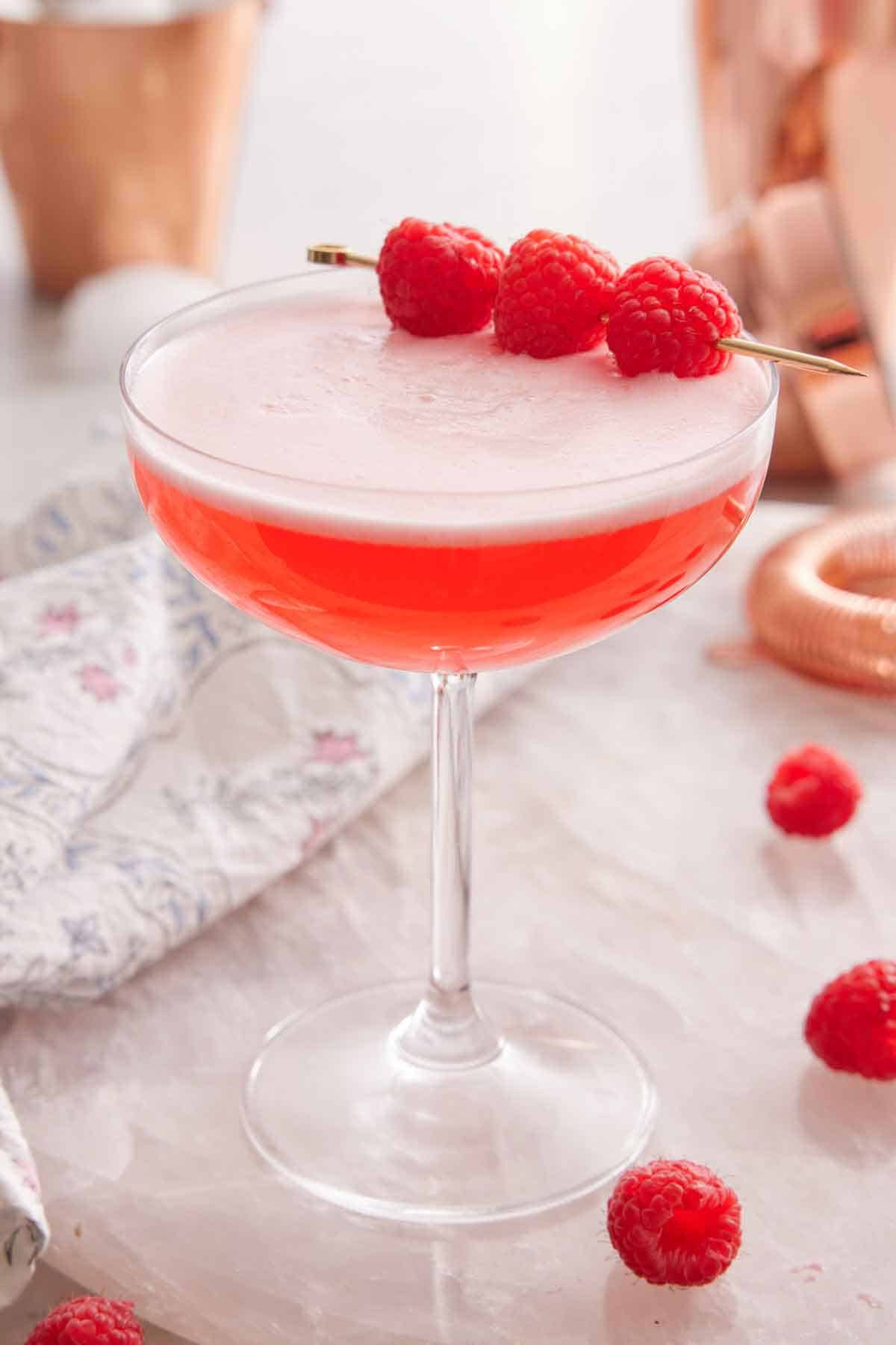 A glass of Clover Club Cocktail with three raspberries as a garnish with more raspberries scattered on the counter.
