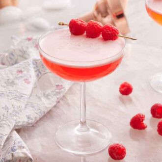 Glass of Clover Club Cocktail with raspberries as a garnish and on the counter.