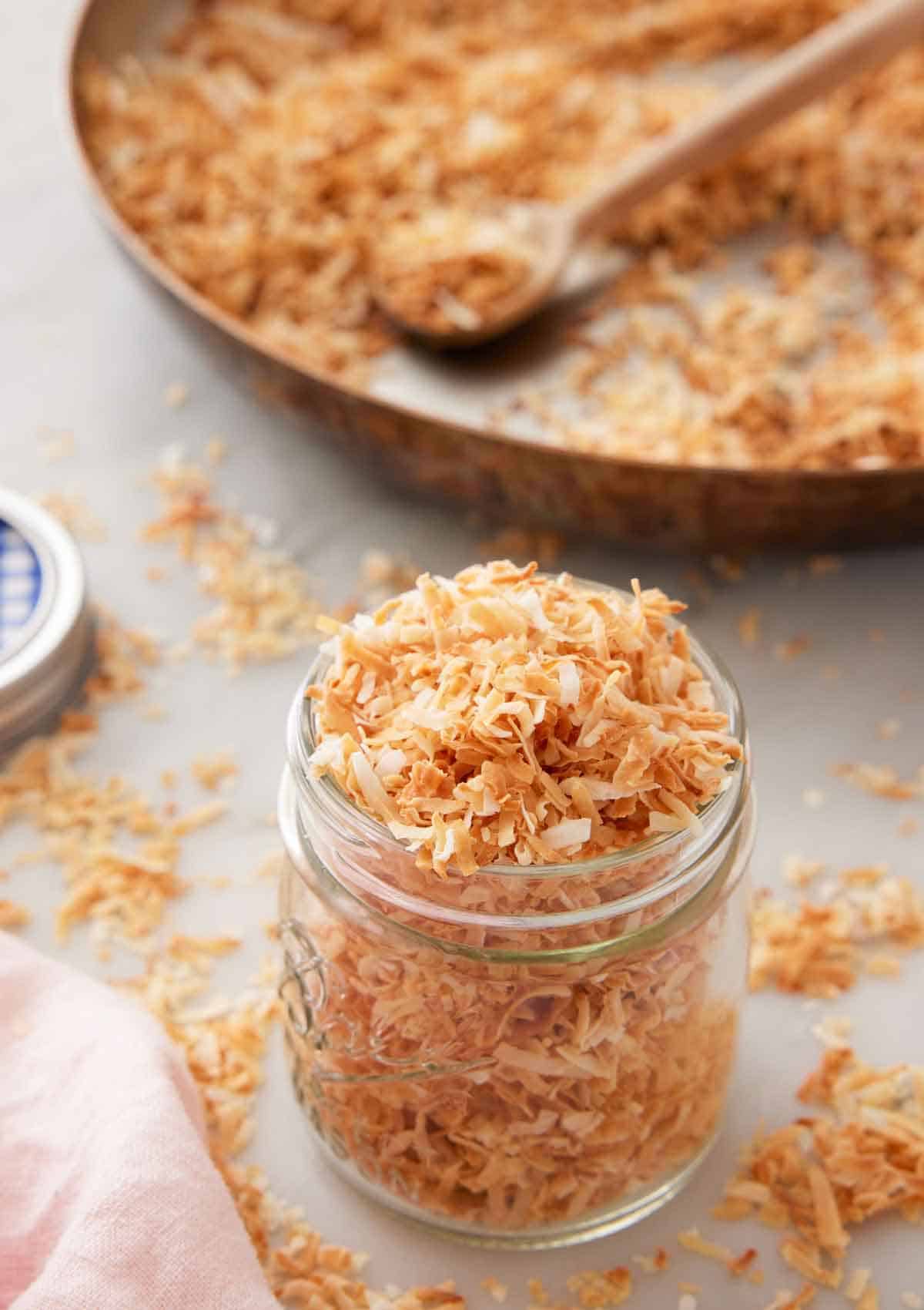 A jar of toasted coconut with a skillet in the background with more coconut.