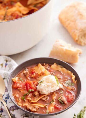 A bowl of lasagna soup with a dollop of cheese in the middle and some torn bread in the background along with a white pot.