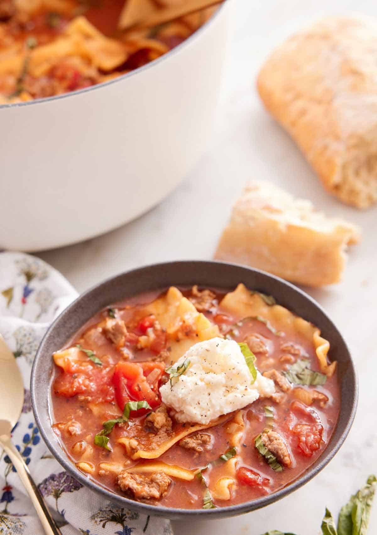A bowl of lasagna soup with a dollop of cheese in the middle and some torn bread in the background along with a white pot.