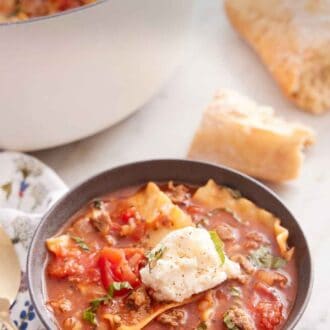 Pinterest graphic of a bowl of lasagna soup with a dollop of cheese in the middle and some torn bread in the background along with a white pot.