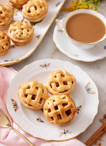 A plate with three mini apple pies with a cup of coffee and platter or more mini apple pies behind it.