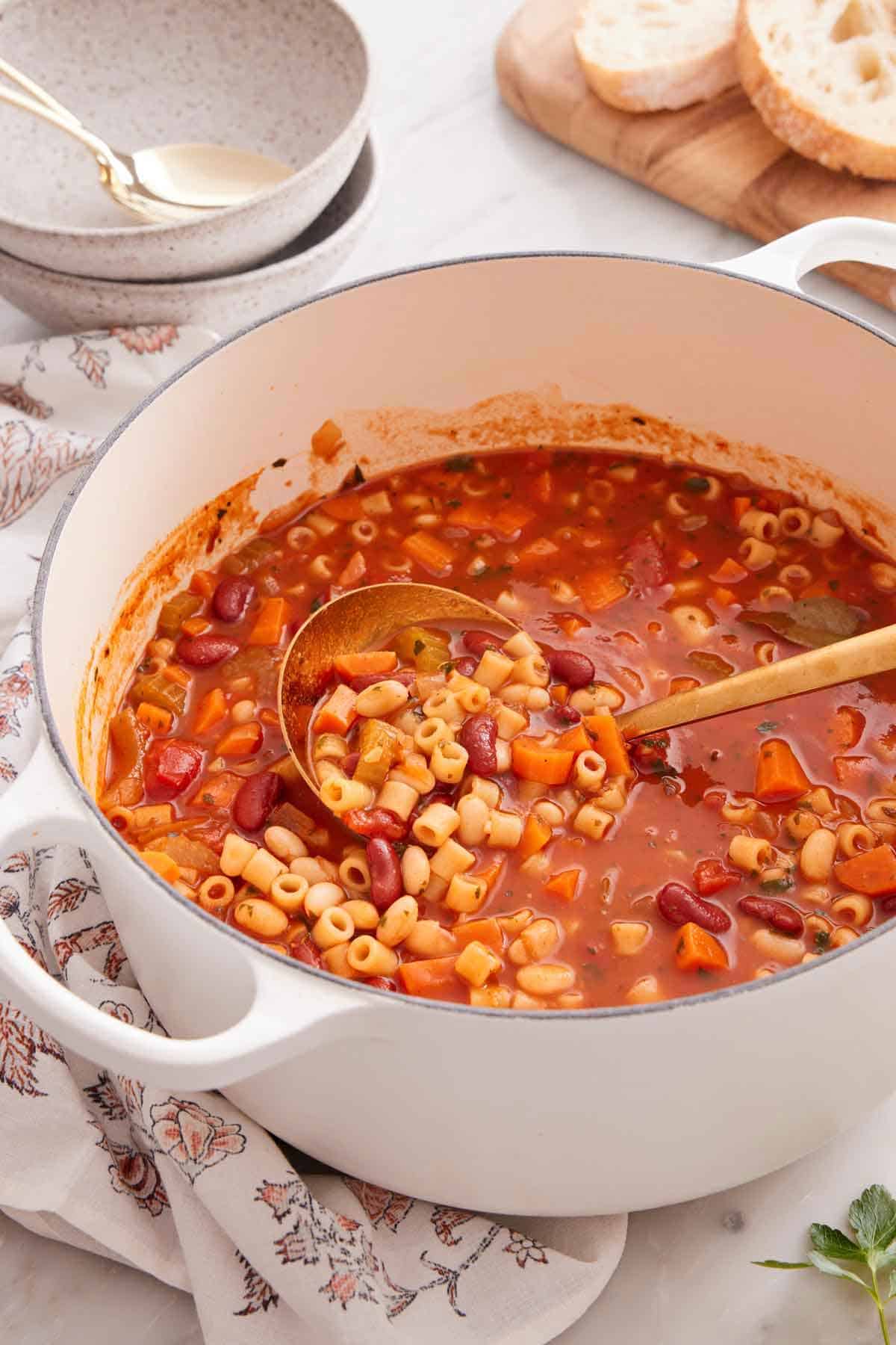A white pot of pasta fagioli with a ladle inside. Bowls and bread in the background.