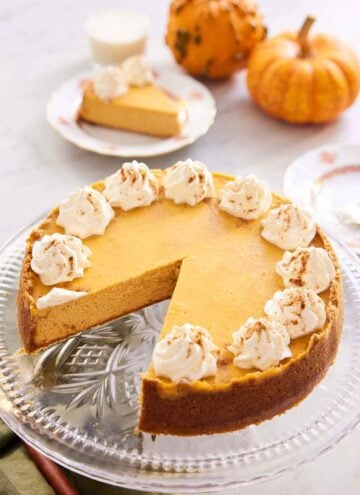 A pumpkin cheesecake on a cake stand with a slice removed. Mini pumpkins and a plated slice in the background.