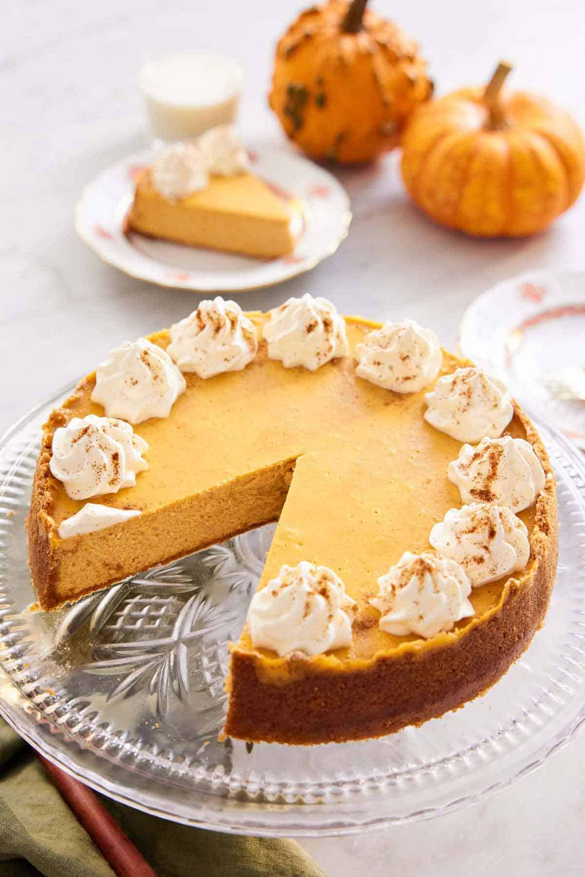 A pumpkin cheesecake on a cake stand with a slice removed. Mini pumpkins and a plated slice in the background.