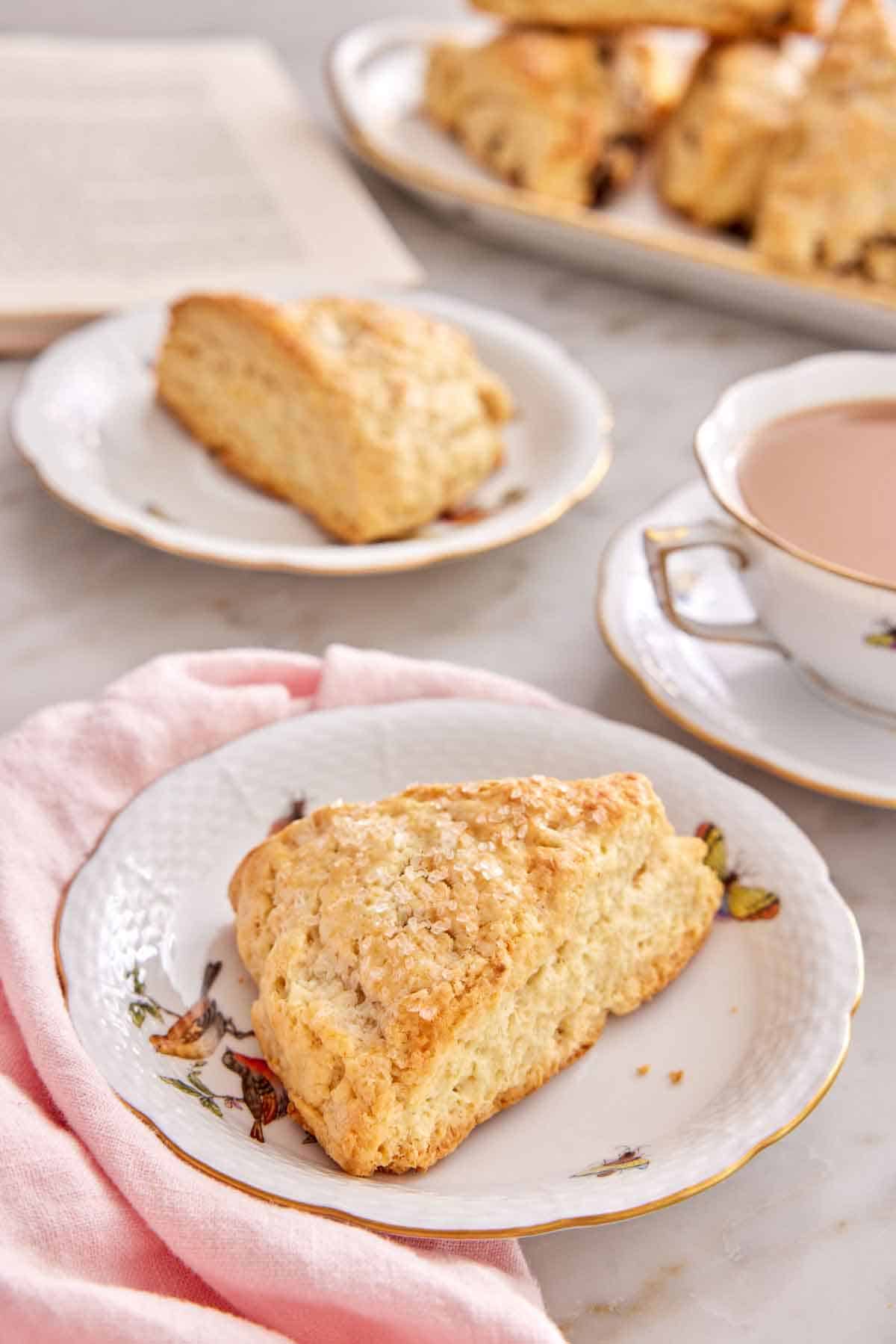 A plate with a scone with a mug of coffee, another plate with a scone, a platter of scones, and an opened book in the background.