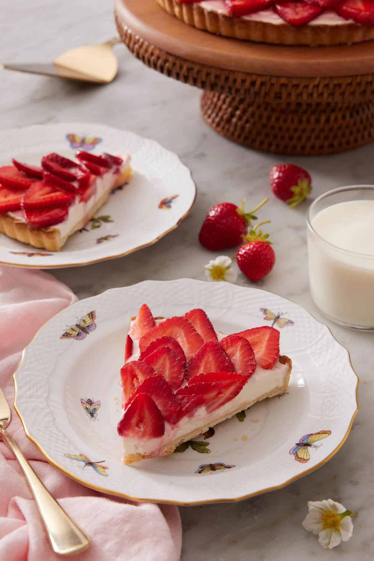 A plate with a slice of strawberry tart with strawberries, glass of milk, a second plated slice, and cake stand in the background.