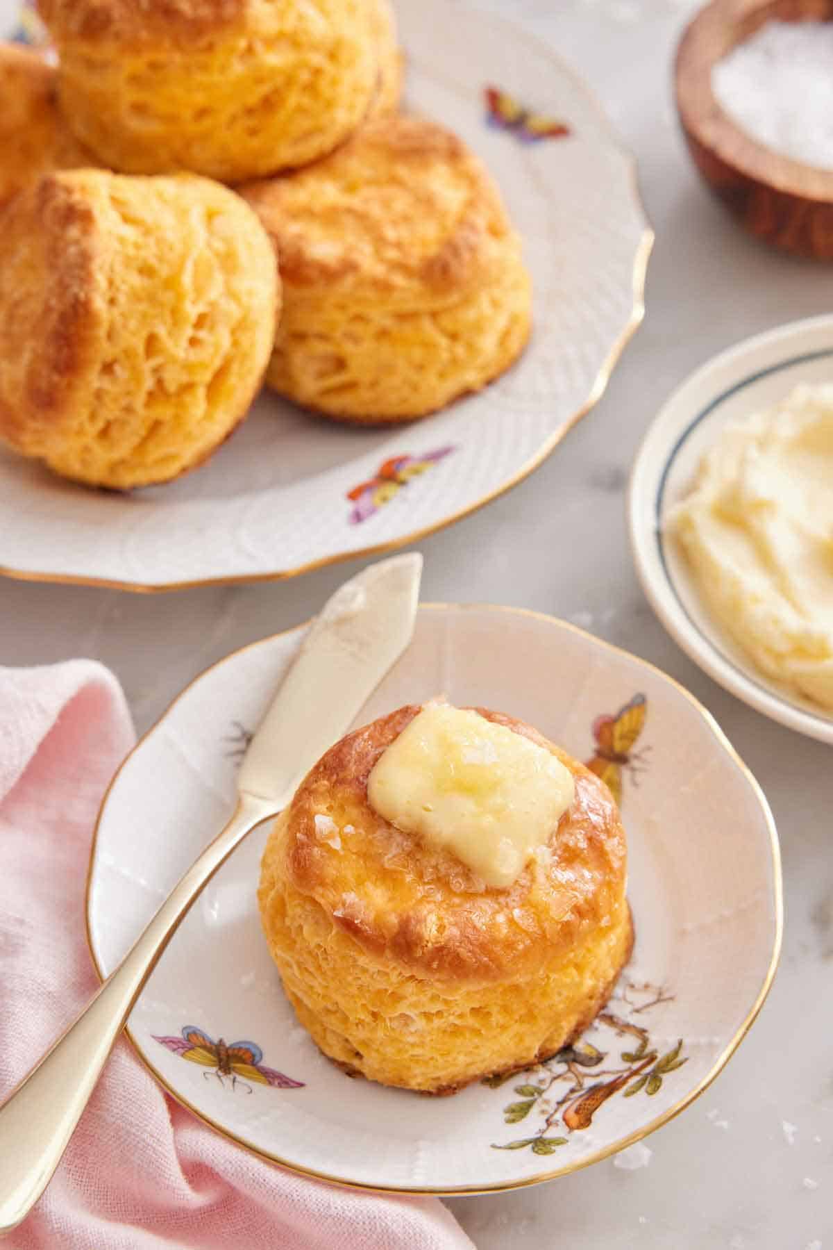 A plate with a sweet potato biscuit topped with butter with additional biscuits on a plate, butter, and salt in the background.