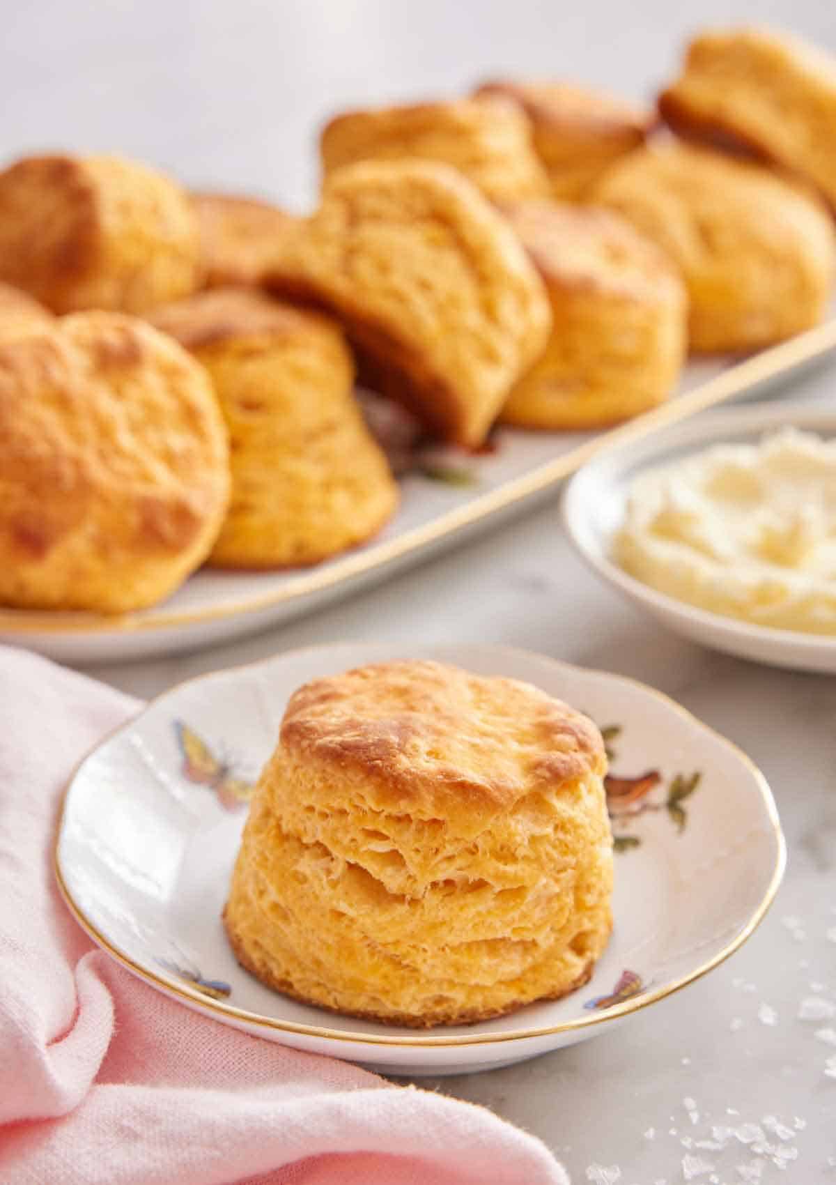A plate with a sweet potato biscuit with a plate of butter and platter of biscuits in the background.