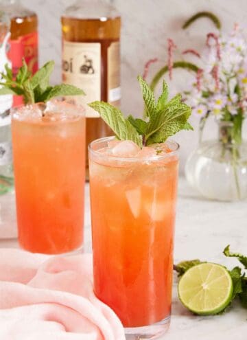 Two glasses of zombie cocktail garnished with mint and a cut lime beside the glasses. Flowers in the background with bottles of alochol.