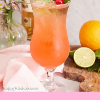 Pinterest graphic of a glass of zombie cocktail garnished with mint leaves and a maraschino cherry. Cut lime and an orange on a cutting board beside it and flowers in the background with a bottle of rum.
