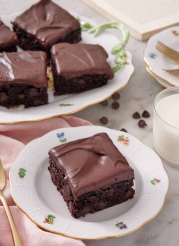 A plate with a slice of zucchini brownie with a platter in the background. Glass of milk on the side.