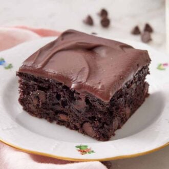 A square piece of zucchini brownie on a plate with a glass of milk and chocolate chips in the background.
