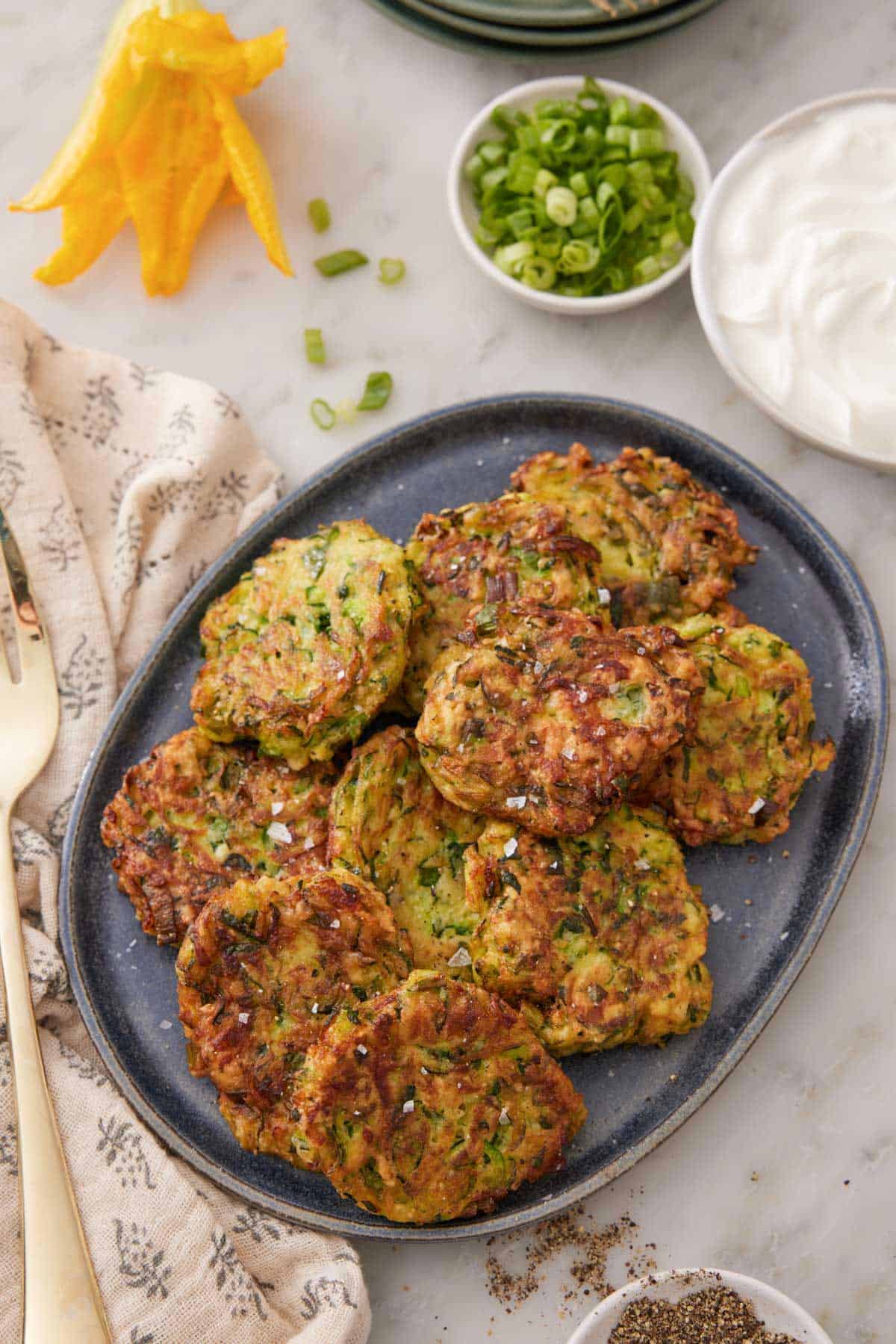 A large platter with a pile of zucchini fritters with green onions, sour cream, and blossom beside it.