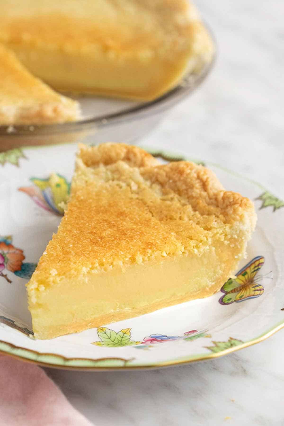 A piece of buttermilk pie on a porcelain plate with the rest of the pie in the background.