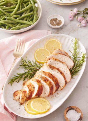 A platter with a sliced air fryer turkey breast on top of rosemary and lemon slices. Green beans in the background.