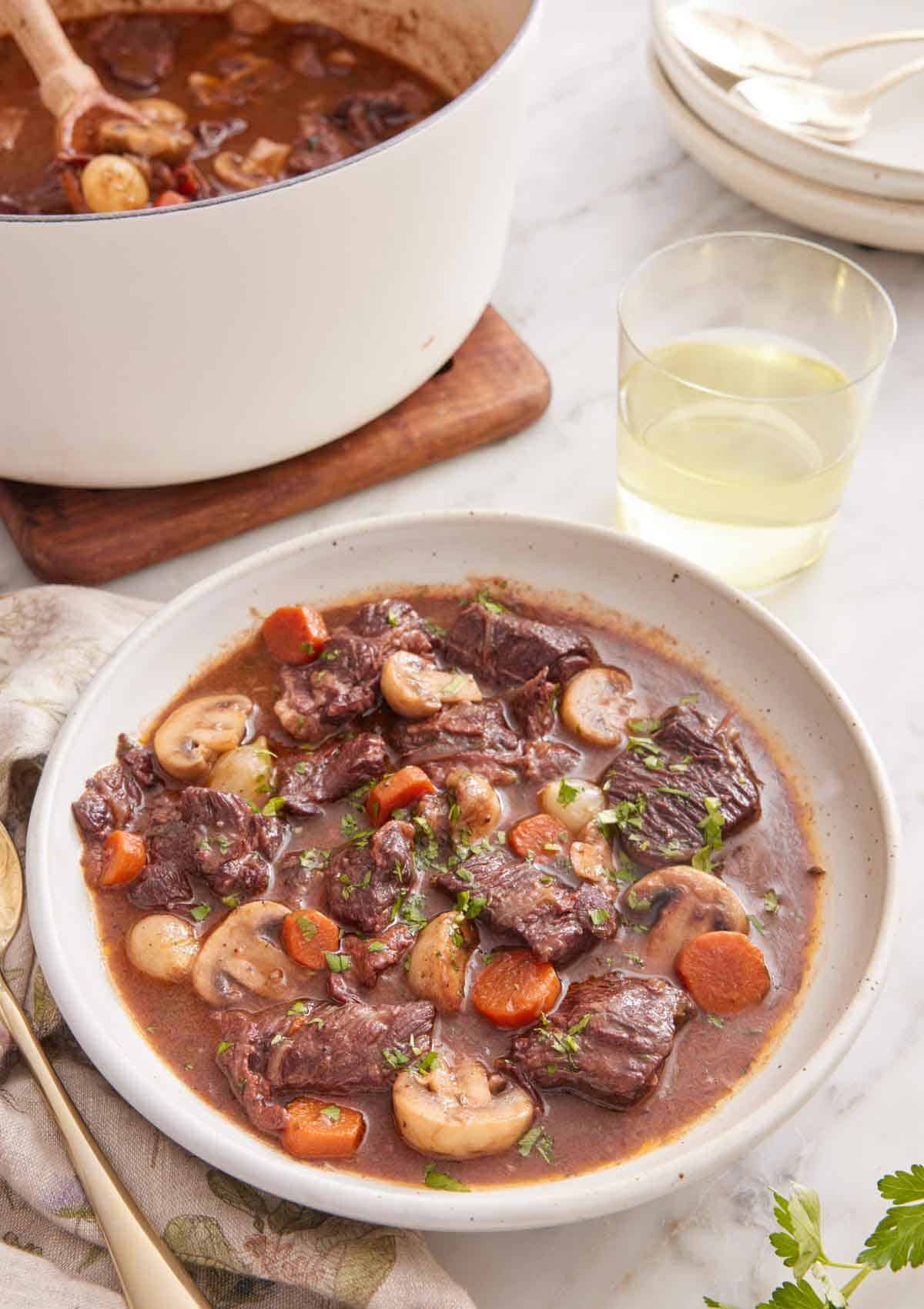A bowl of beef bourguignon with a glass of wine and pot of more beef bourguignon in the background.