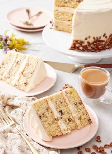 Two plated slices of butter pecan cake with the rest of the cake on a cake stand in the background with a glass of coffee.