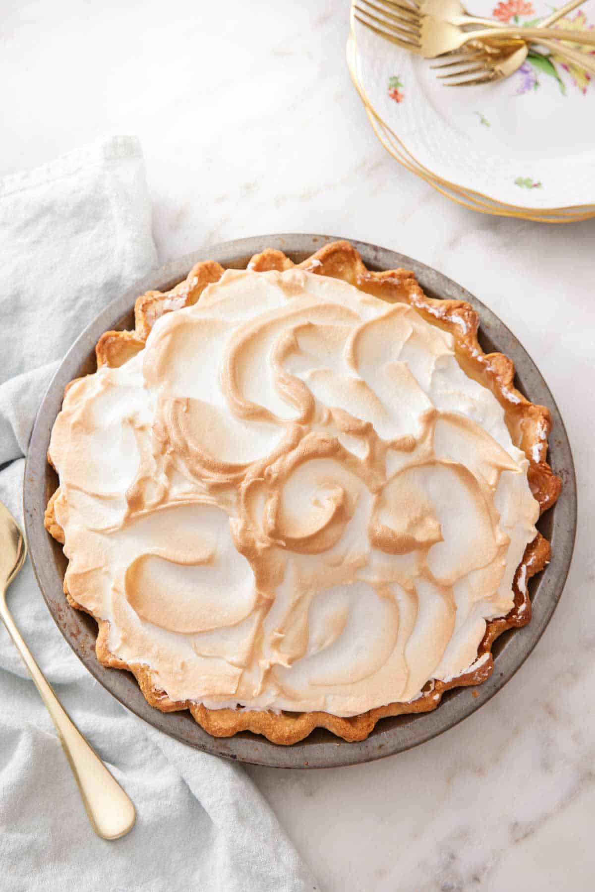 A slightly overhead view of a butterscotch pie in its baking dish. Stack of plates and forks on the side.