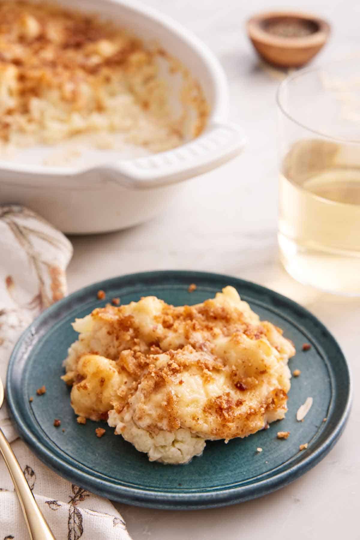 A plate with a serving of cauliflower gratin with a baking dish in the background along with a glass of wine.