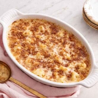 A white baking dish containing a cauliflower gratin. A serving spoon and stack of plates beside it.
