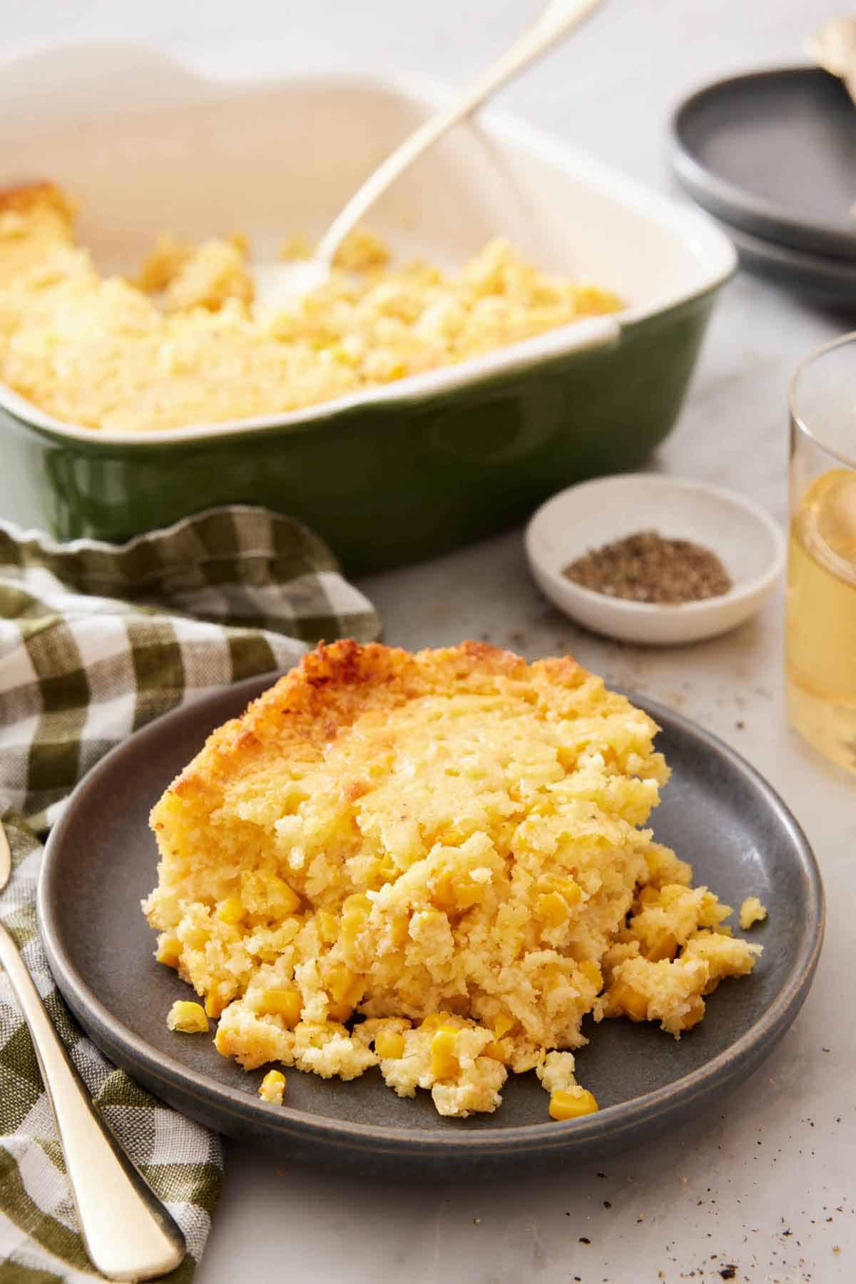 A plate with a serving of corn casserole with a baking dish, pepper, and drink in the background.