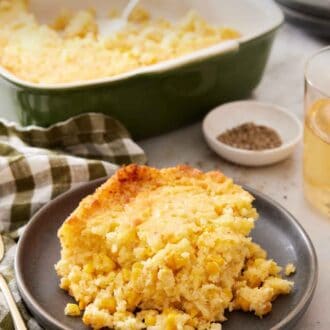 Pinterest graphic of a plate with a serving of corn casserole with a baking dish, pepper, and drink in the background.