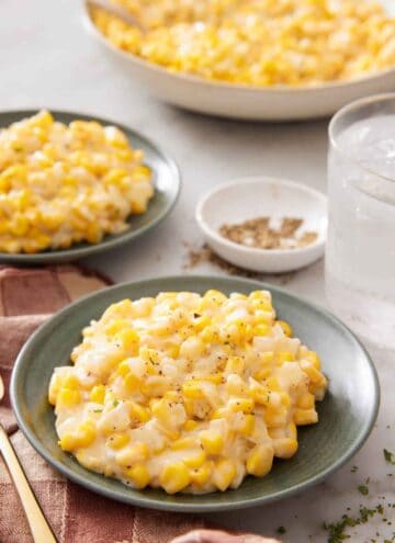 Two plates of creamed corn with a platter with more in the background along with a glass of water and bowl of pepper.