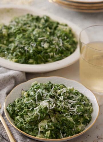 A bowl of creamed spinach with a drink and serving platter of creamed spinach in the background.