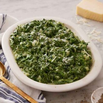 A serving platter of creamed spinach with a block of grated parmesan, bowl of pepper, and a serving spoon beside it.