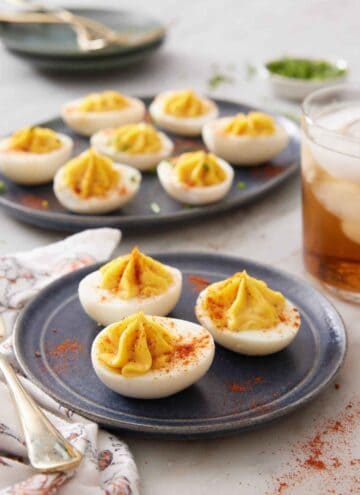A plate with three deviled eggs with an additional platter in the background with more. A drink off to the side and paprika sprinkled around.