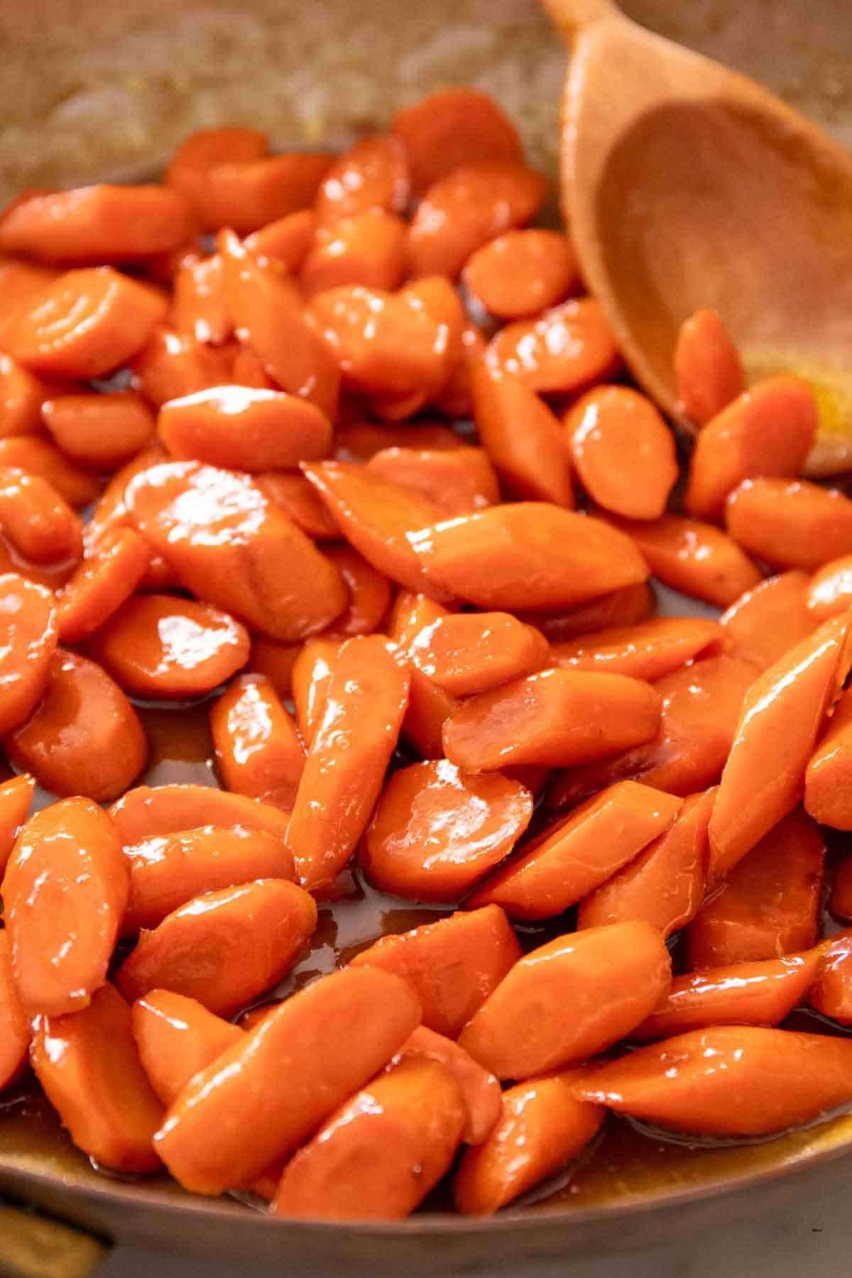 A skillet of glazed carrots with a wooden spoon in the background.